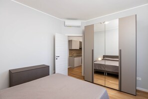 Un dormitori modern amb llit, una taula de nit, un armari i una cuina visible a través d'una porta oberta.