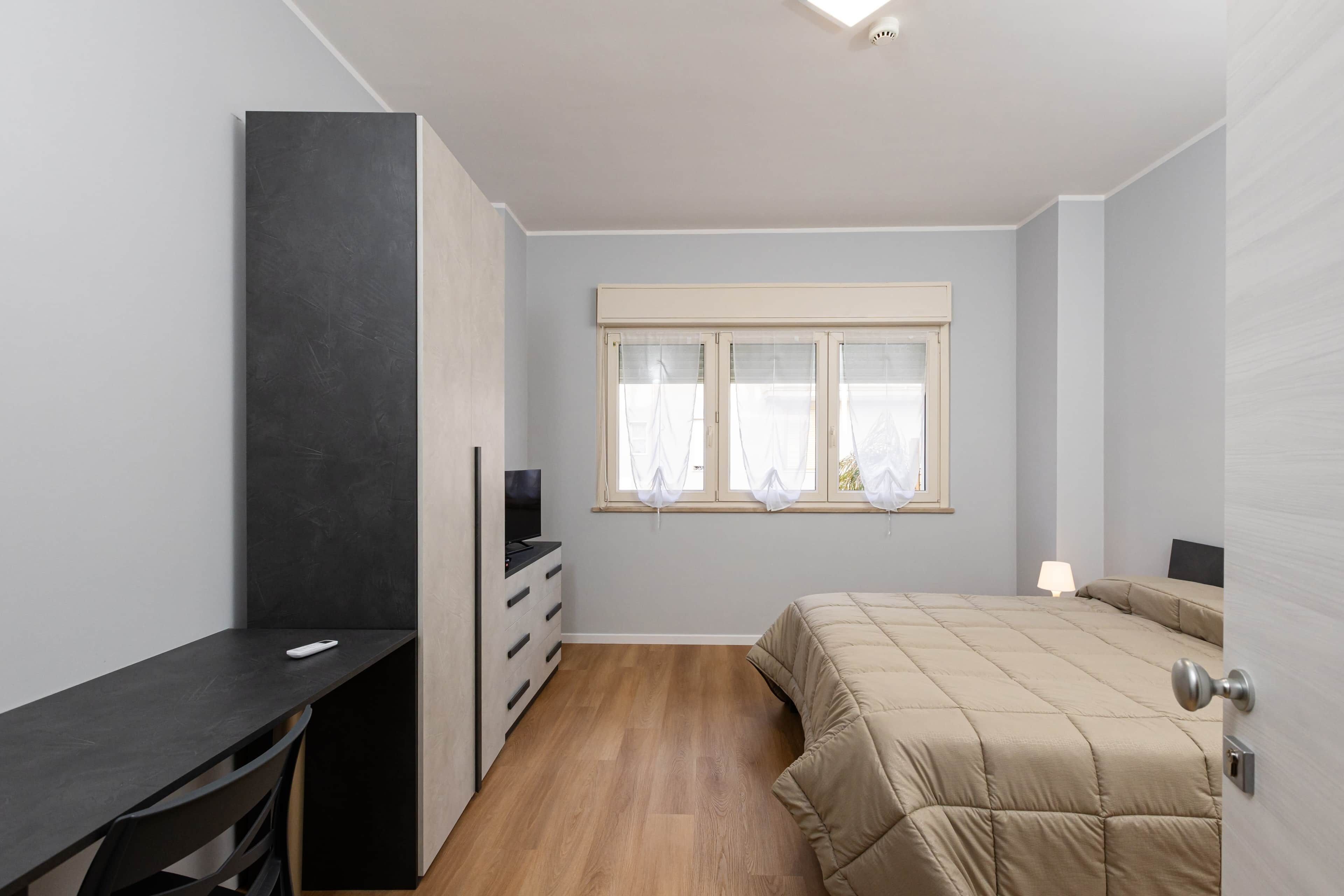 Un dormitorio con cama, escritorio, televisor y ventana con cortinas.
