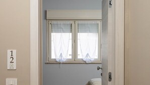 Ein schmaler Flur, der in ein Schlafzimmer mit Bett, Fenster mit Vorhängen und Tür führt.