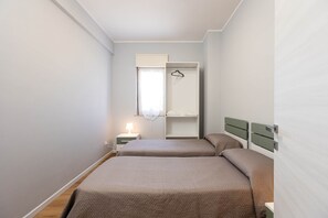 Una habitación pequeña y bien iluminada con dos camas, una mesita de noche con lámpara, una ventana con cortina y un perchero.