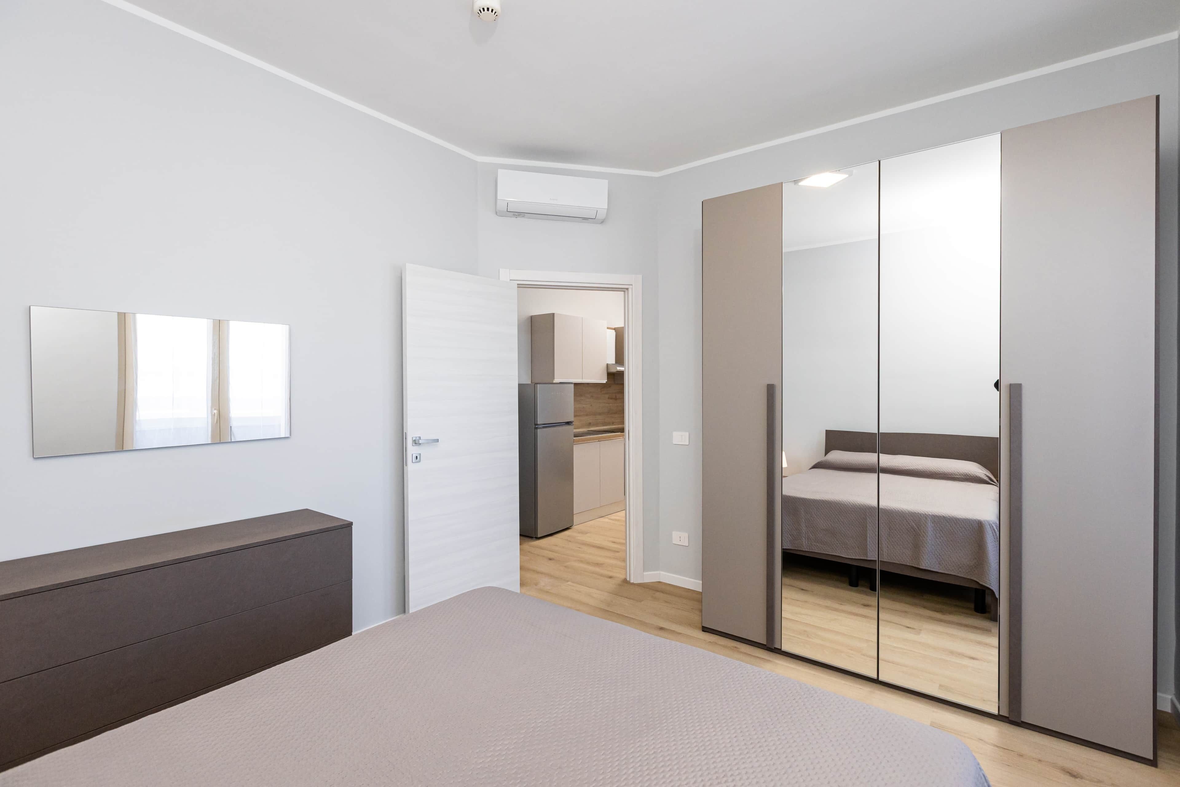 A modern bedroom with a bed, a dresser, a wardrobe, and a kitchen in the background.