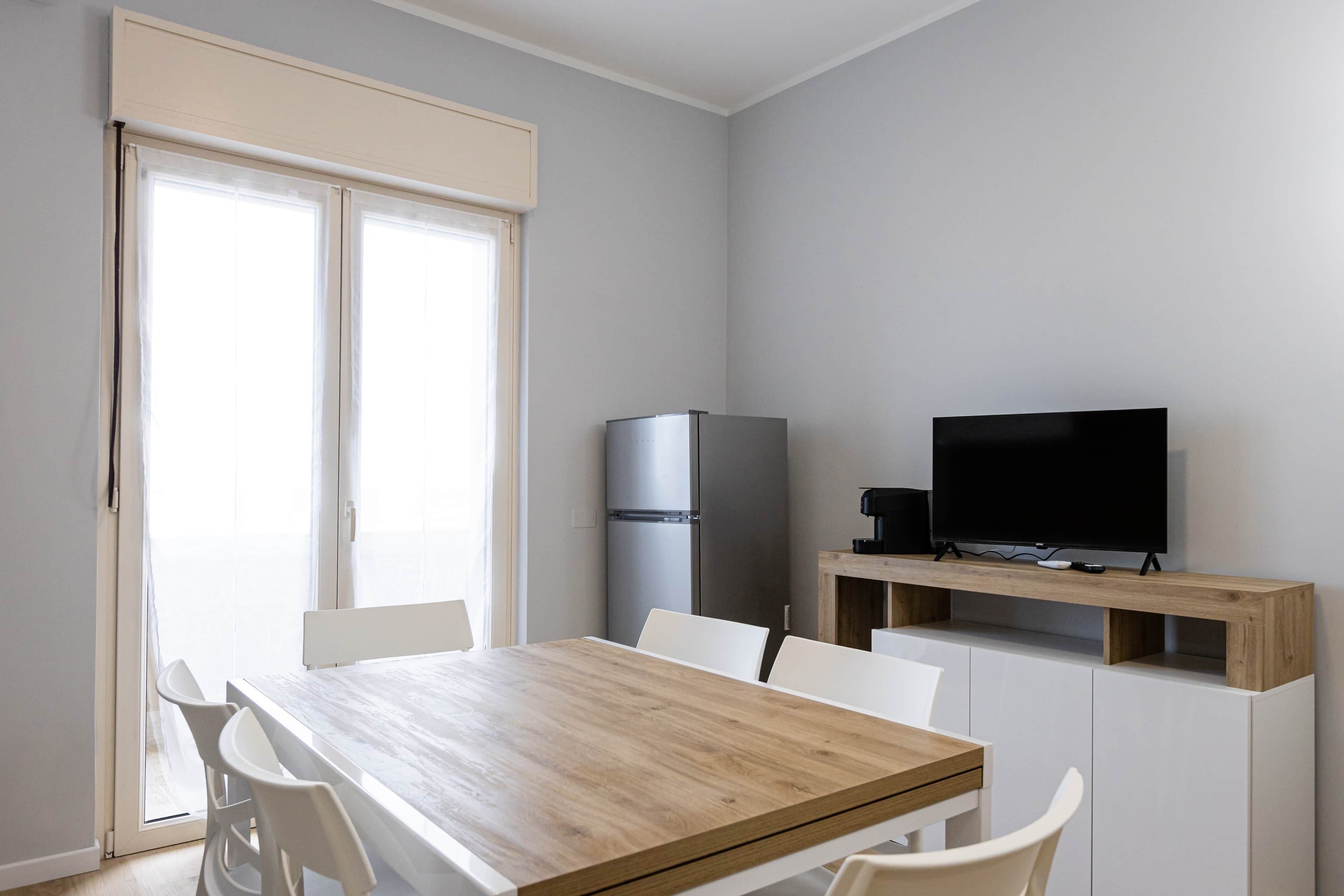 A modern kitchen with a wooden table, white chairs, a refrigerator, and a TV on a wooden shelf.