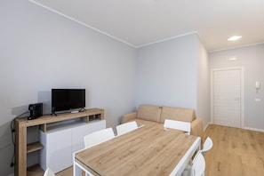 Una habitación moderna y luminosa con una mesa de comedor de madera, sillas blancas, un sofá beige, un televisor de pantalla plana sobre un mueble de madera y una puerta blanca.