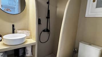 A compact bathroom with a white sink, a round mirror, a handheld showerhead, a toilet, and a trash bin.