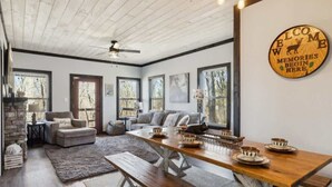 A spacious living room with a wooden dining table set for four, a fireplace, and a large window with a view of trees. - Sassy Bear Hideaway by Beyond Expectations: 4BR w/ Firepit, Game Room & Hot Tub (Sevierville)