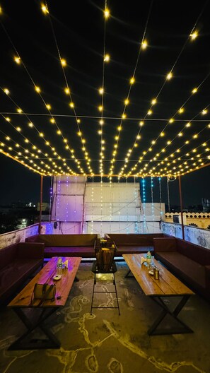 Un espace extérieur aménagé avec des tables et des bancs, des guirlandes lumineuses suspendues au-dessus, et une vue sur le paysage urbain en arrière-plan.