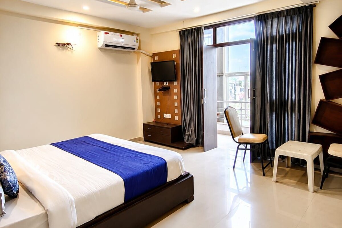 A modern hotel room with a bed, a desk, a chair, a flat-screen TV, and an air conditioning unit.