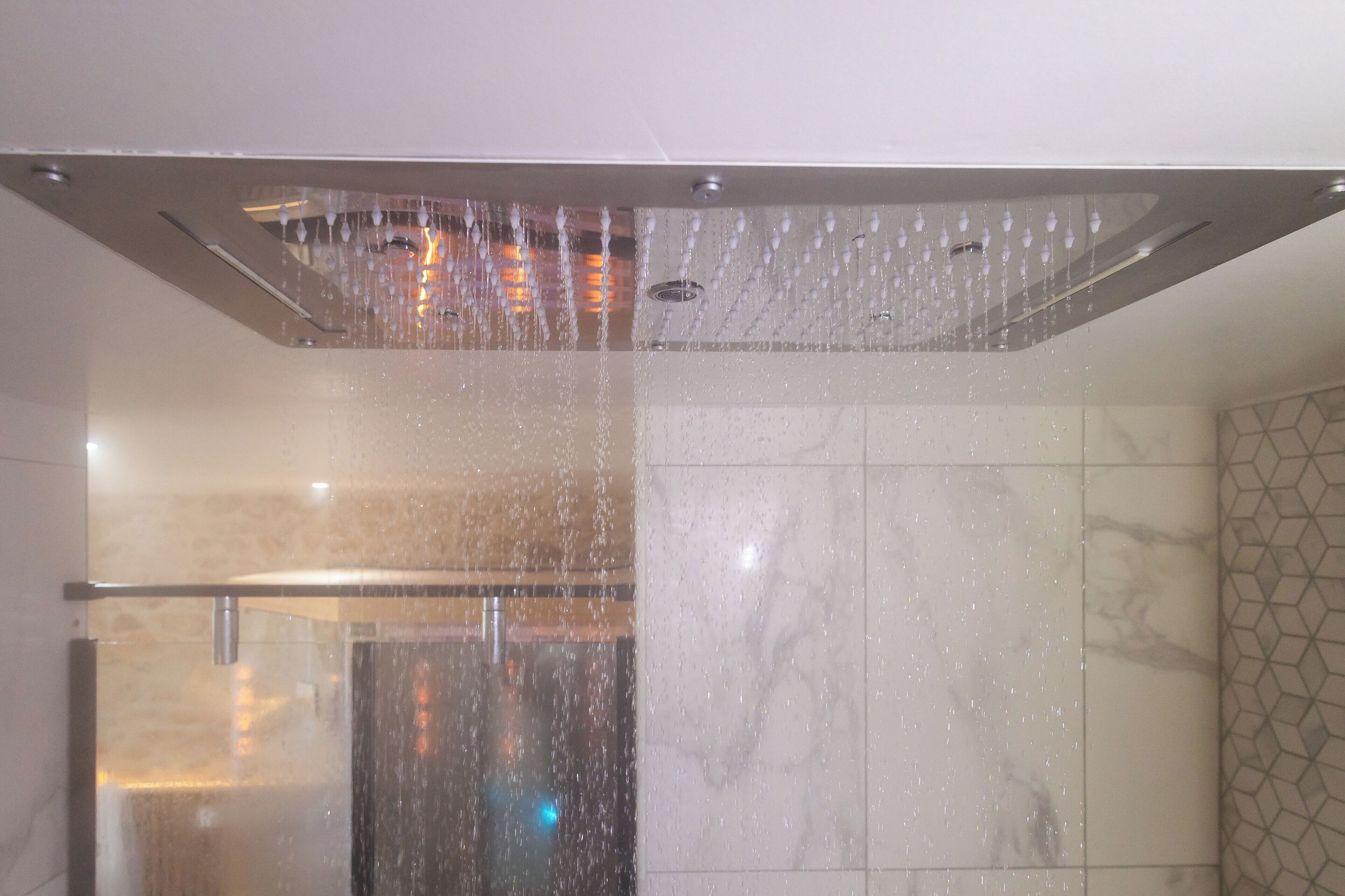 Une douche dotée d’un pommeau de douche créant un effet de pluie, d’une paroi en verre et d’un mur carrelé orné d’un motif géométrique.