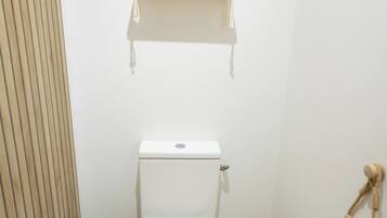 A small, clean bathroom with a toilet, a trash bin, a shelf with a potted plant, and a wooden slat wall.