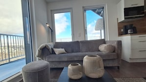 A modern living room with a grey sofa, two windows, a coffee machine, and a view of the city. - Spacious Apartment with Exceptional Mountain View, Sauna and Terrace (Winterberg)