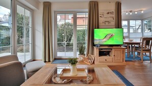 Ett modernt vardagsrum med en stor TV, möbler i trä och utsikt genom stora fönster.