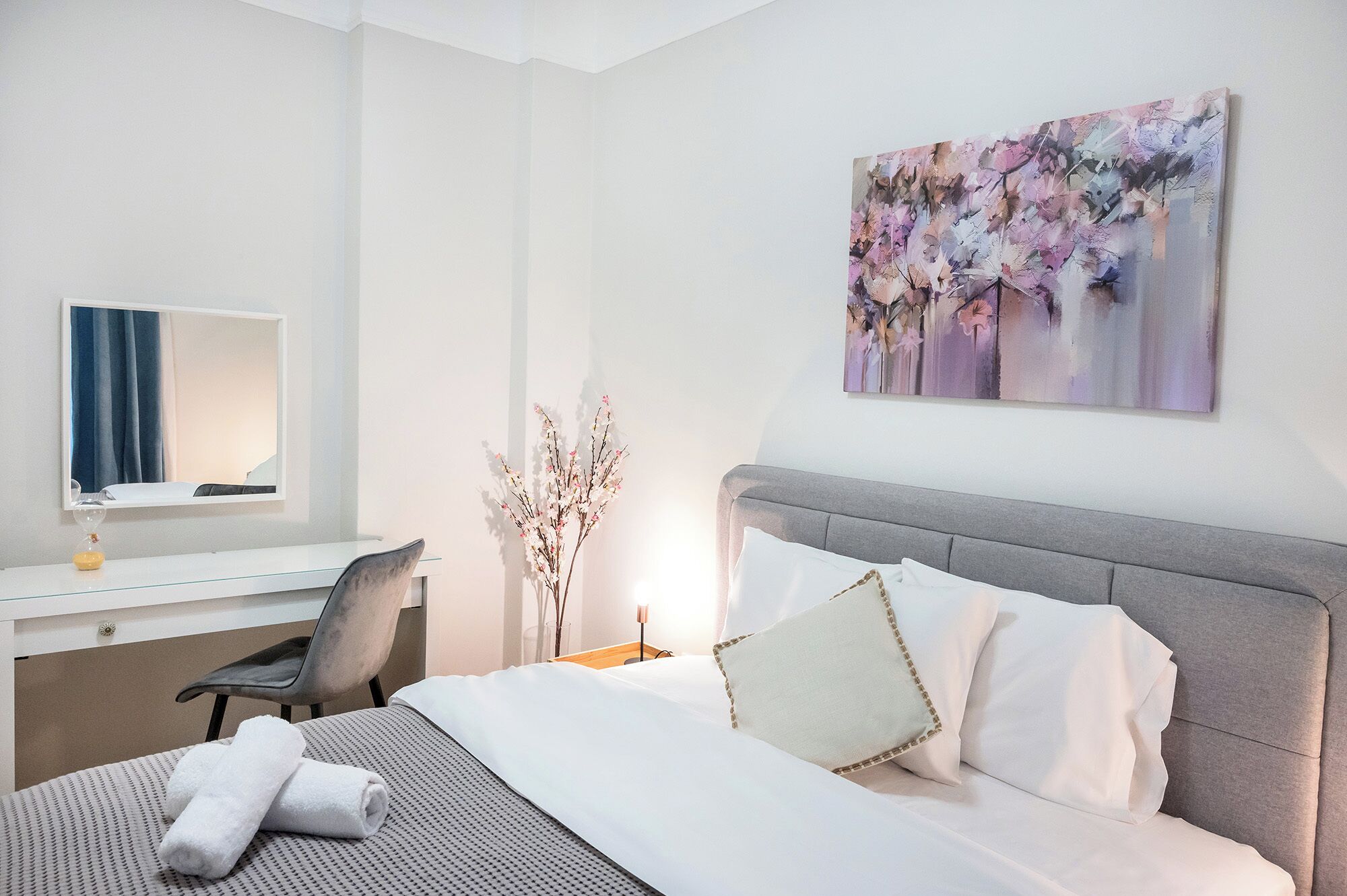 A hotel room with a bed, a desk, a chair, a mirror, and a wall-mounted painting.
