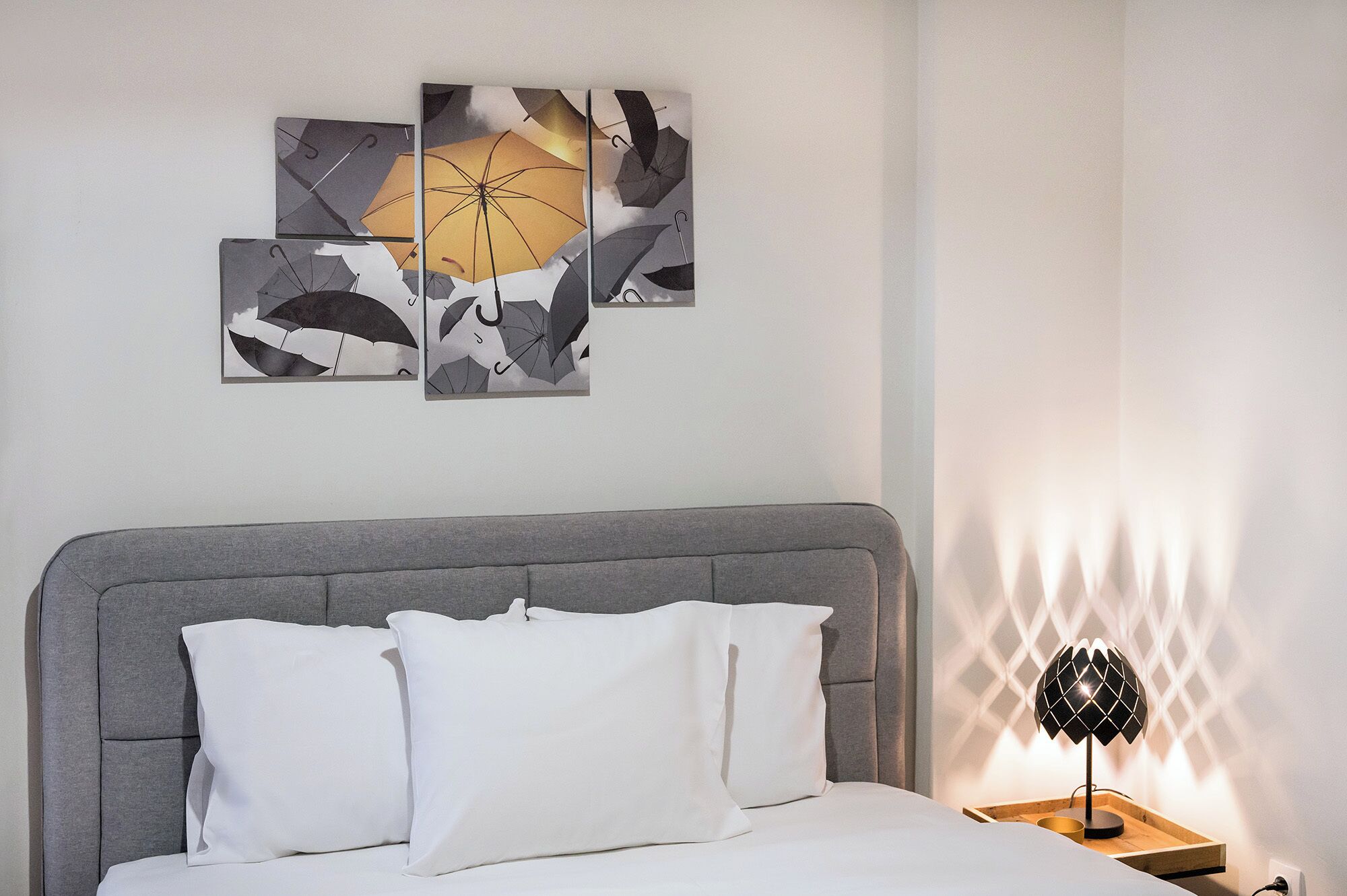 A bedroom with a grey upholstered headboard, white bedding, and a wall-mounted artwork of abstract umbrellas.