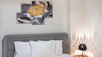 A bedroom with a grey upholstered headboard, white bedding, and a wall-mounted artwork of abstract umbrellas.