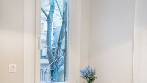Een klein raam met een witte lijst, met uitzicht op een boom en gebouwen. Daaronder een witte radiator, en op de tafel een vaas met blauwe bloemen.