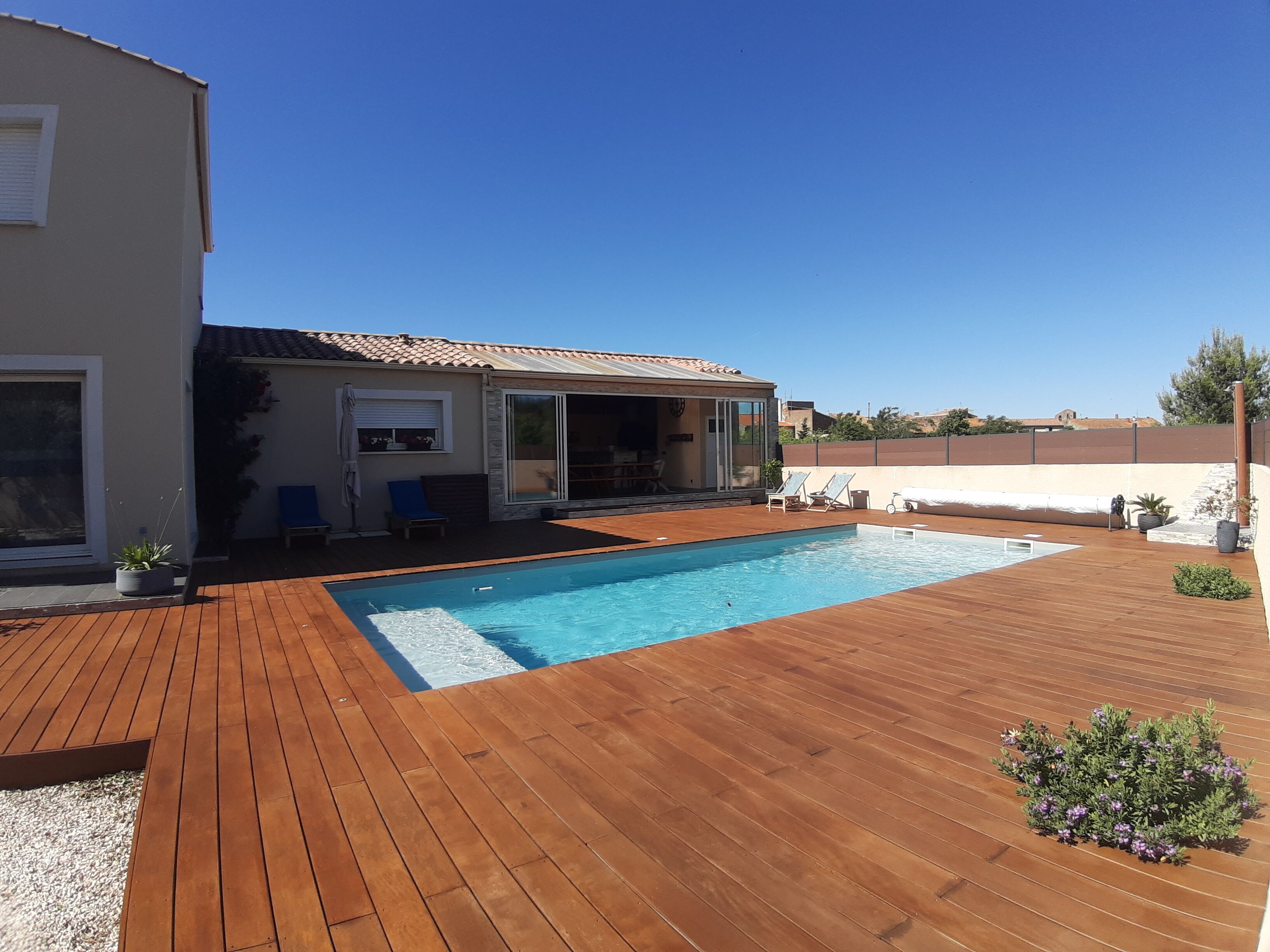 Ein Schwimmbad mit Holzterrasse, einer überdachten Terrasse und einem Haus mit Glastüren.