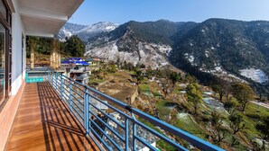 A balcony with a wooden floor and blue railings overlooking a mountainous landscape with snow-capped peaks and a river.