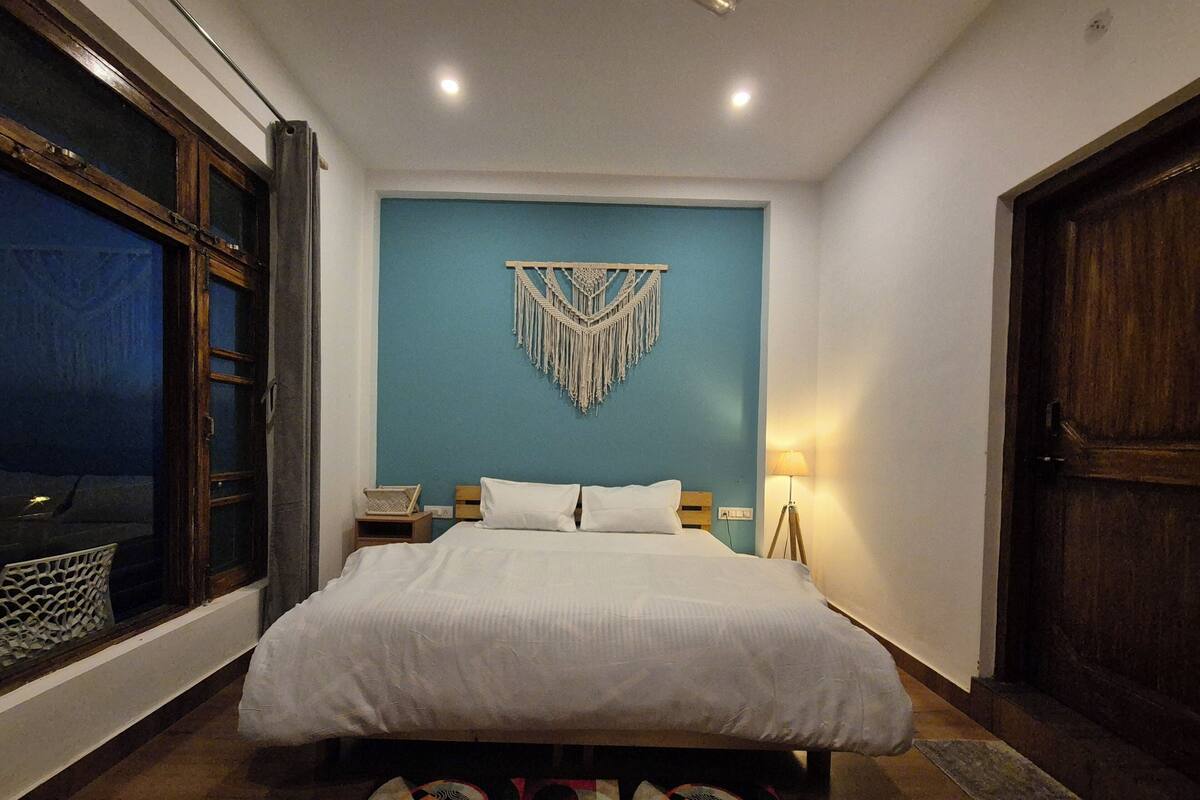 A bedroom with a bed, a macrame wall hanging, a bedside lamp, and a view through a window.