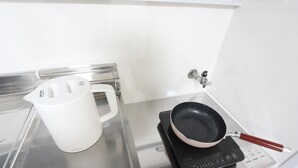 Een keuken met een witte waterkoker, een koekenpan op een draagbaar fornuis en een roestvrijstalen aanrecht.