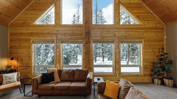 A log cabin interior with a high ceiling, large windows, a brown leather sofa, and a small table.