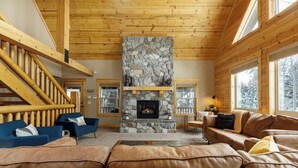 A cozy living room with a stone fireplace, wooden beams, and large windows.