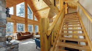 A log cabin interior with a wooden staircase, stone fireplace, and large windows offering views of snow-covered trees.