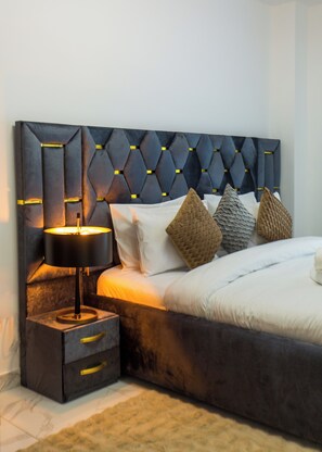 A modern bedroom with a tufted headboard, a bedside table with drawers, and a lamp. - Premium Two Bedroom Suite at Essence (Accra)