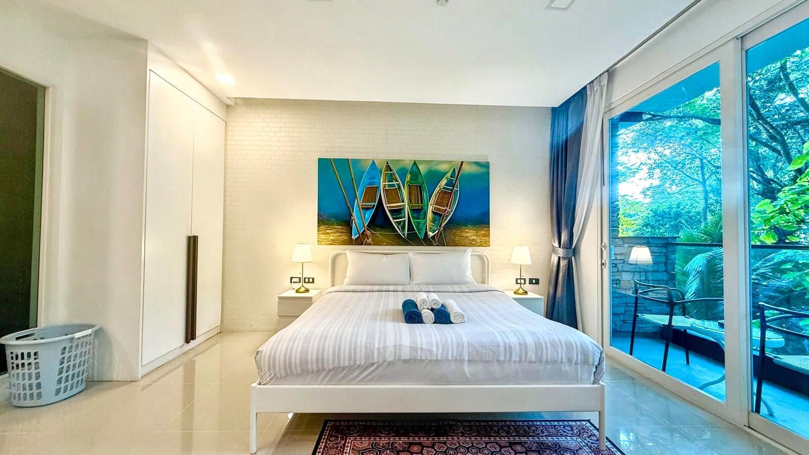 A modern bedroom with a bed, two bedside tables, a painting, and a view of greenery through large windows.