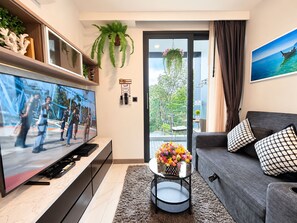 Un soggiorno moderno con una TV a schermo piatto, un divano grigio, un tavolino rotondo con un vaso di fiori e un'ampia finestra vista su del verde.