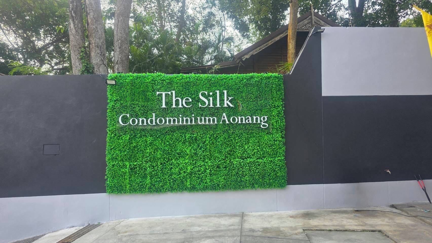 A sign for "The Silk Condominium Aonang" with a green hedge backdrop.
