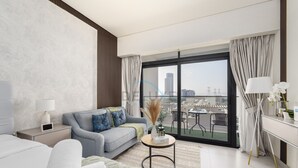 Een moderne hotelkamer met een grijze bank, een klein rond tafeltje met een lamp, een nachtkastje met een lamp, en een groot raam met uitzicht op de stad.
