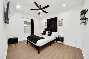 Een moderne slaapkamer met een bed, een plafondventilator, een nachtkastje, een zwart voetenbankje en een aan de muur bevestigde televisie.