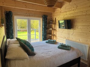 Een houten slaapkamer met een bed, een televisie en uitzicht op een groen landschap door een schuifdeur.