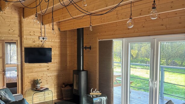 A cozy cabin interior with a sofa, a TV, a fireplace, and a large sliding glass door to the outdoors. - 3 Bedroom 2 Bath Chalet with hot tub in peaceful countryside close to Exeter (Broadclyst)