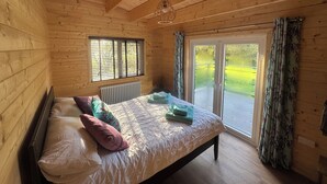A wooden cabin bedroom with a bed, window, and sliding glass door to a balcony. - 3 Bedroom 2 Bath Chalet with hot tub in peaceful countryside close to Exeter (Broadclyst)