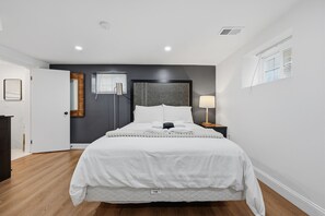 A modern bedroom with a large bed, a bedside table with a lamp, and a door to the left. - Near DC Group Getaway Free Parking + WiFi + Yard (Hyattsville)
