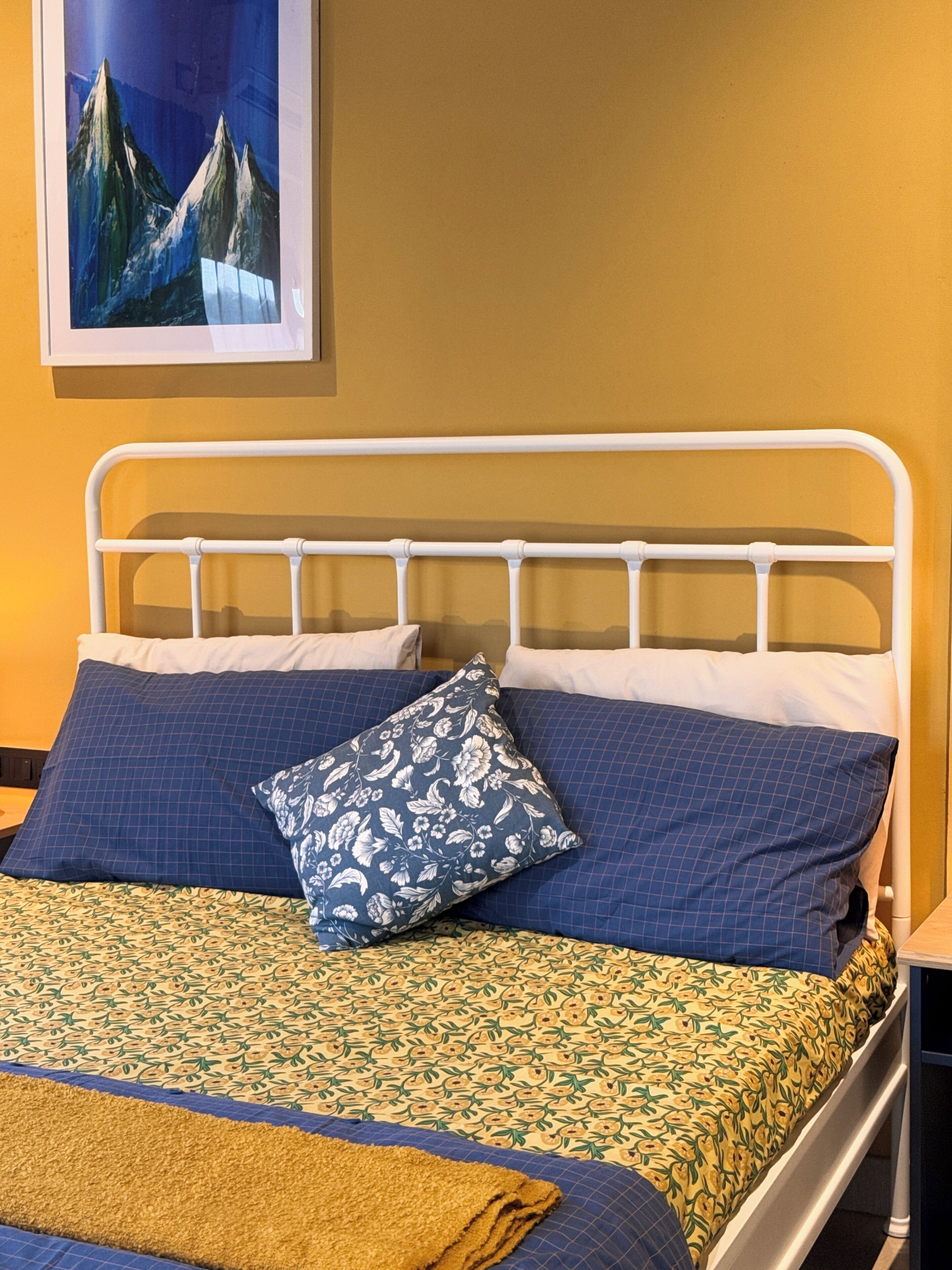 Una cama con estructura metálica blanca, ropa de cama azul estampada y una manta amarilla.