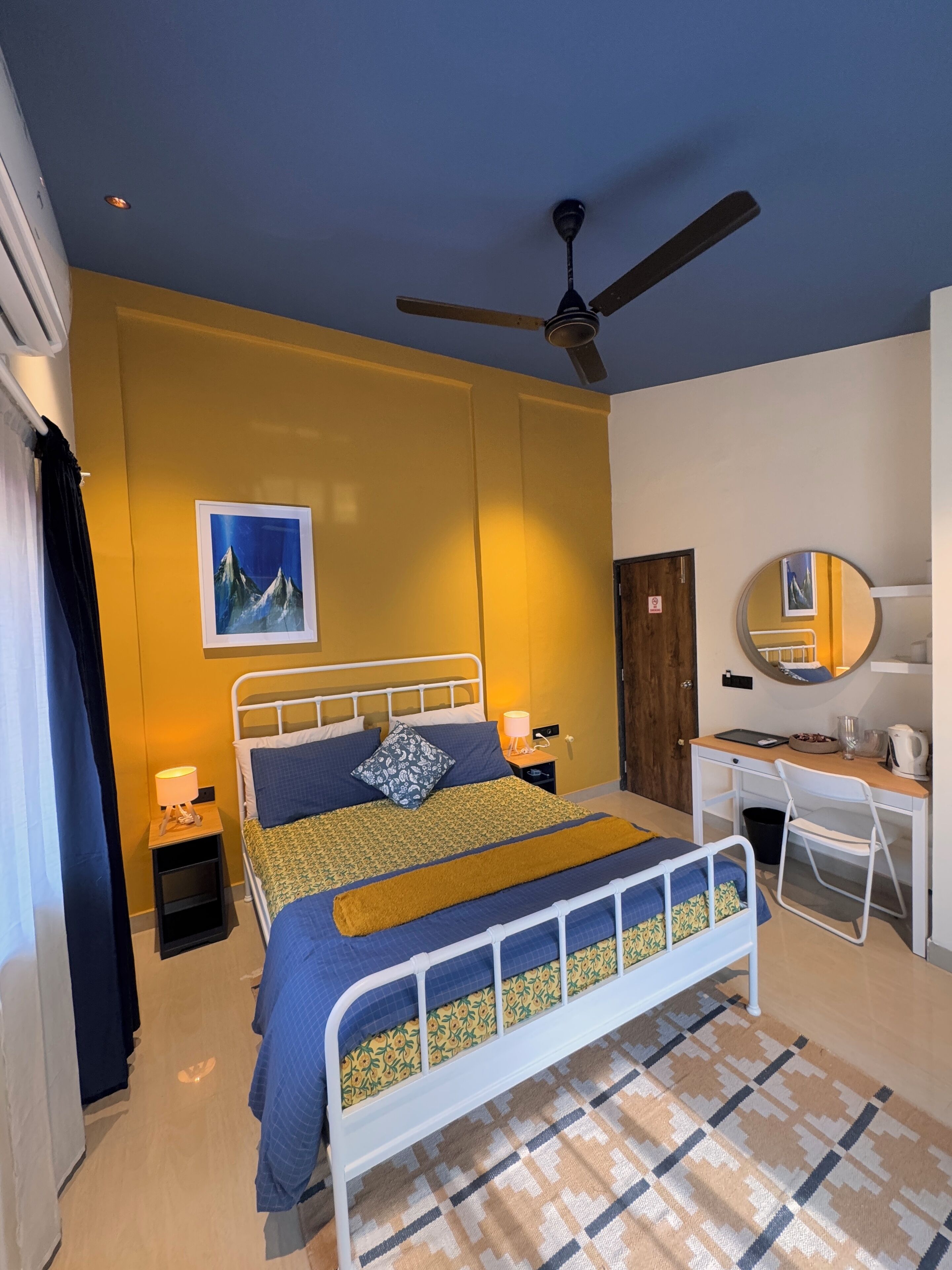 A bedroom with a bed, bedside tables, a desk, a chair, a ceiling fan, a mirror, and a picture on the wall.