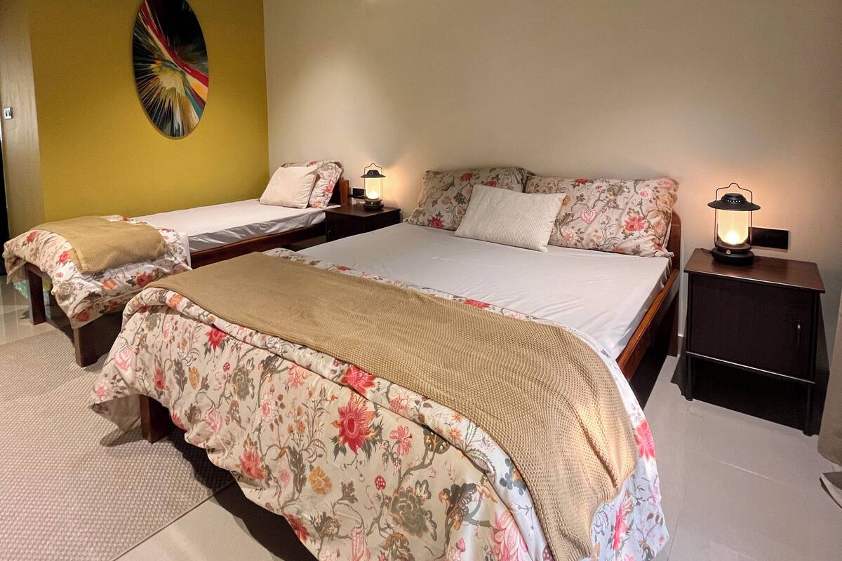 A bedroom with a large bed, two smaller beds, a ceiling fan, an air conditioning unit, and a wall-mounted lamp.