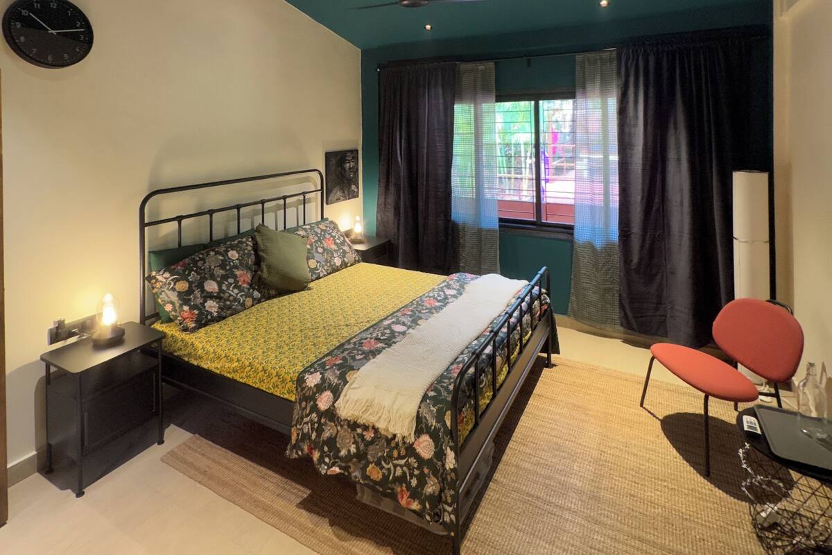 A bedroom with a bed, a nightstand, a clock, a ceiling fan, and a window with curtains.
