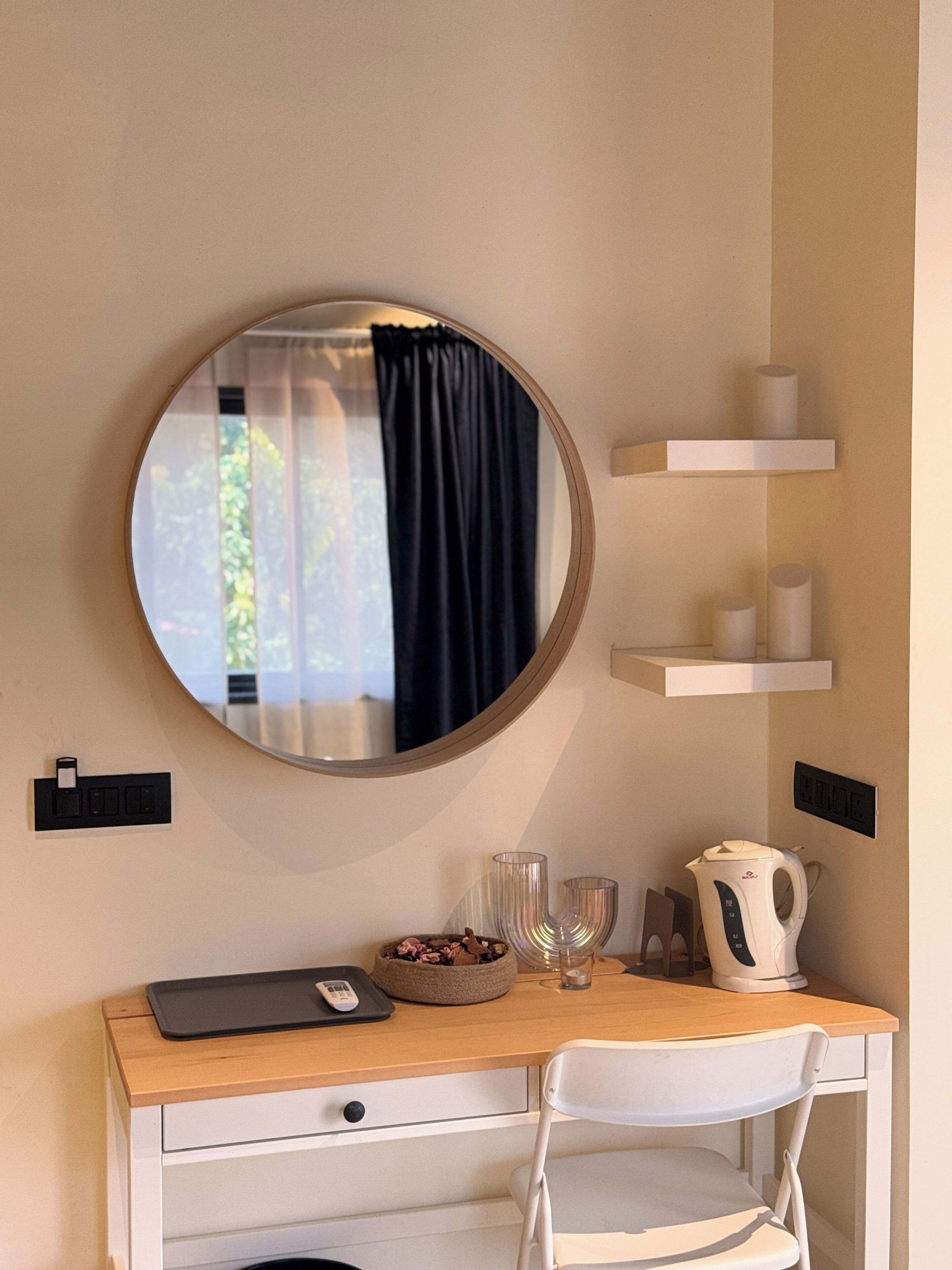 A room with a round mirror, a desk with a chair, a bowl of fruit, a kettle, and a window with curtains.