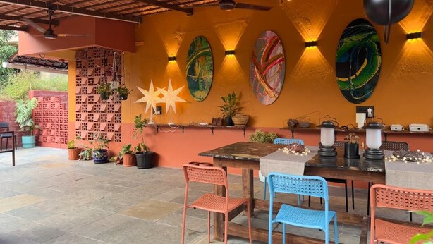 An outdoor patio with a wooden table, blue and red chairs, decorative wall art, and potted plants.