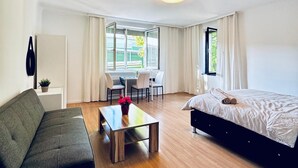A modern hotel room with a bed, a sofa, a dining table with chairs, and a window with curtains. - Spacious Garden Villa for 16 Guests (Vienna)