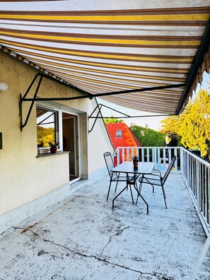 A balcony with a table and chairs, a striped awning, and a view of a red-roofed building. - Spacious Garden Villa for 16 Guests (Vienna)