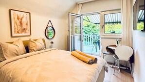 A bedroom with a large bed, a small table, and a chair, a window with a view of greenery, and a ceiling light. - Spacious Garden Villa for 16 Guests (Vienna)