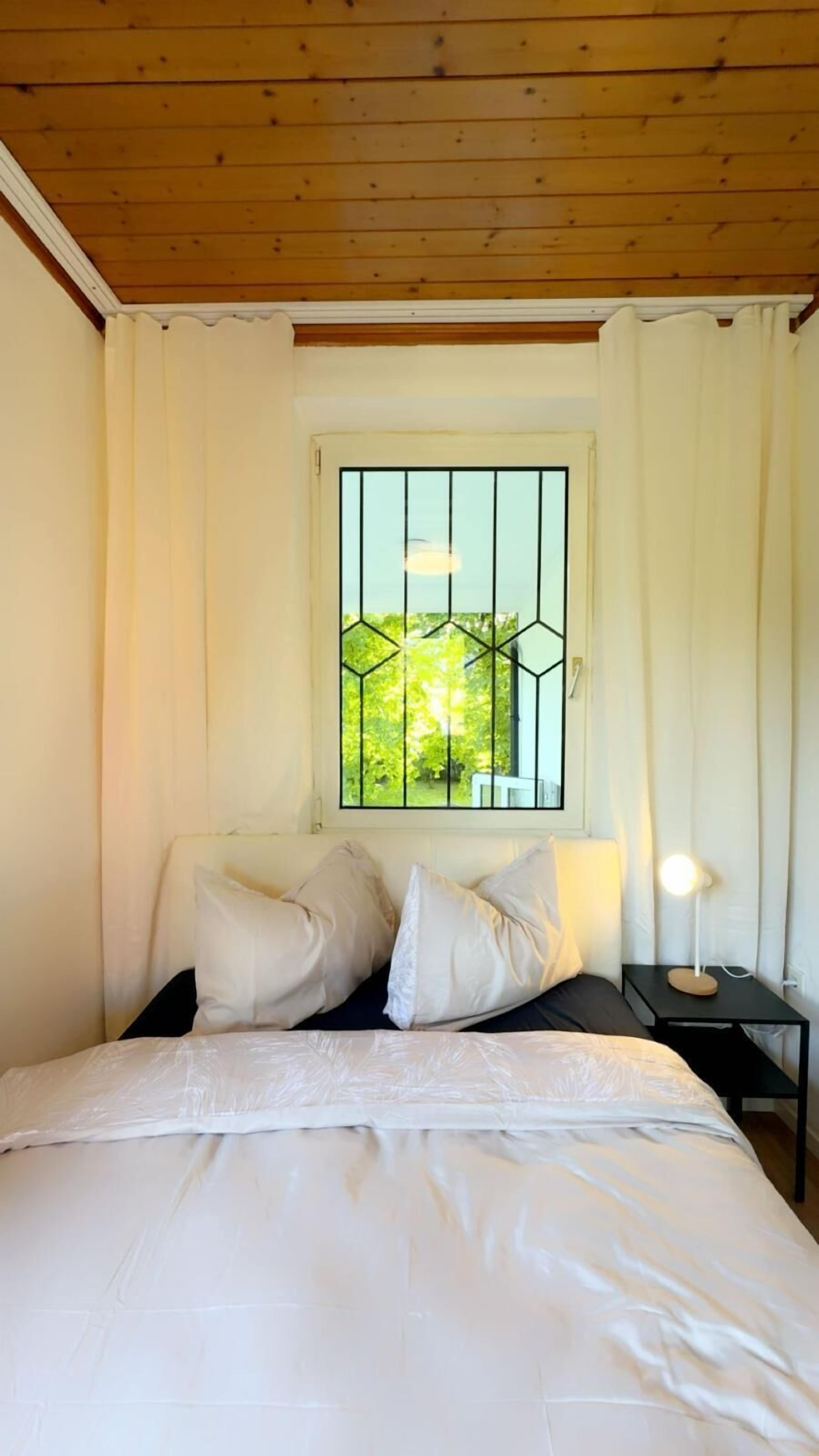 A bedroom with a bed, pillows, a nightstand, a lamp, and a window with a view of greenery.