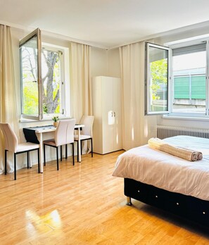 Une chambre à coucher avec un lit, un bureau et une chaise. Il y a des fenêtres avec des rideaux, un radiateur et une armoire.