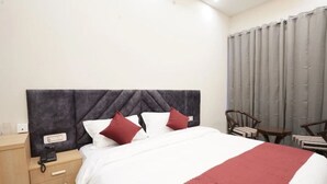 A hotel room with a bed, a nightstand, a chair, and a window with curtains. - Su Casa Banquets and Hotel (Vikasnagar)