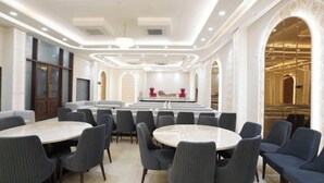 A spacious conference room with round tables and chairs, a stage with red chairs, and a large arched doorway. - Su Casa Banquets and Hotel (Vikasnagar)