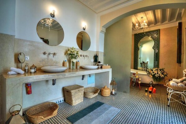 Une salle de bain avec deux lavabos, de grands miroirs et une baignoire. On y trouve des paniers en osier et des bougies sur le sol.